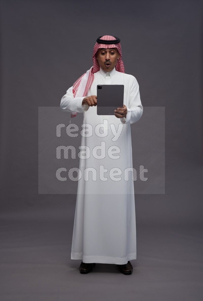 Saudi man wearing thob and shomag standing working on tablet on gray background