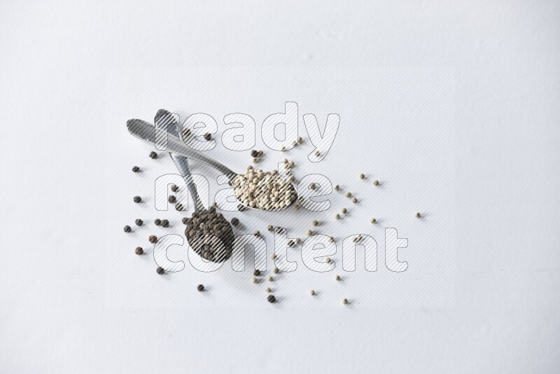 2 metal spoons full of black and white pepper beads with spreaded beads on white flooring