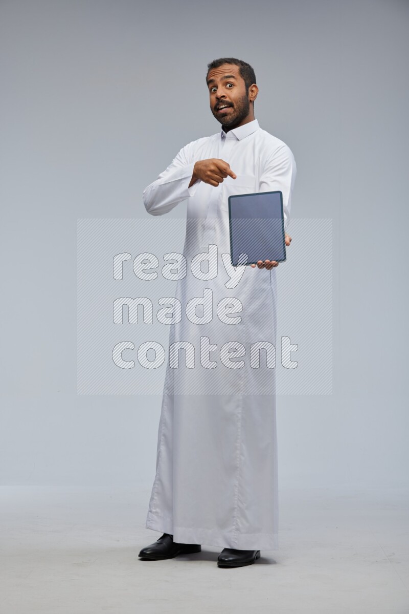 Saudi man wearing Thob standing showing tablet to camera on Gray background