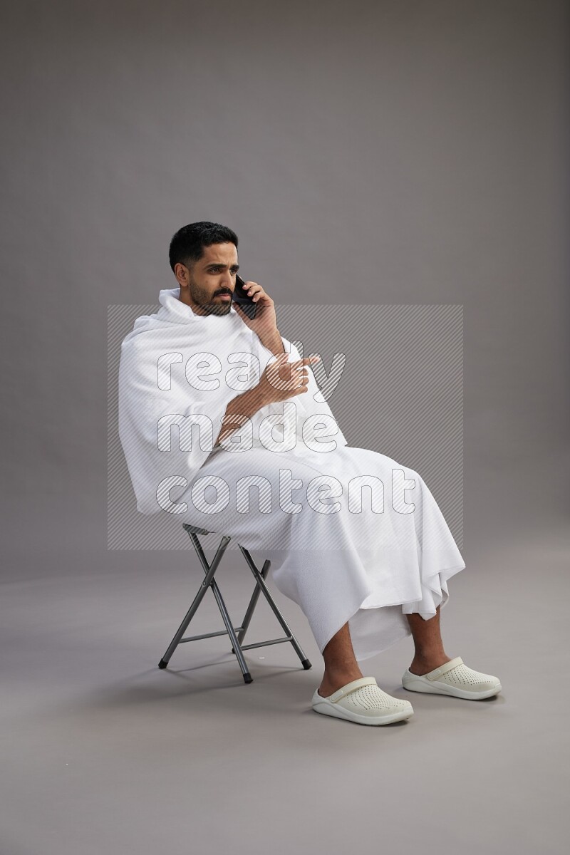 A man wearing Ehram sitting on chair talking on the phone on gray background