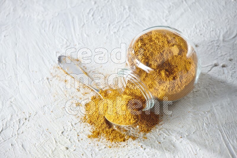 A flipped glass spice jar and a metal spoon full of turmeric powder and powder spilled out of it on textured white flooring