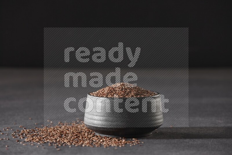 A black pottery bowl full of flaxseeds surrounded by the seeds on a black flooring