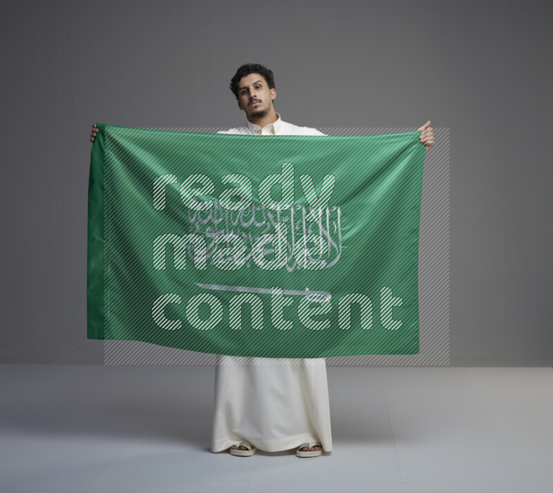 A Saudi man standing wearing thob holding big Saudi flag on gray background