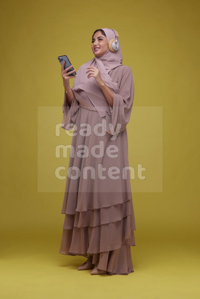 A woman Listening to Music on a Yellow Background wearing Brown Abaya with Hijab