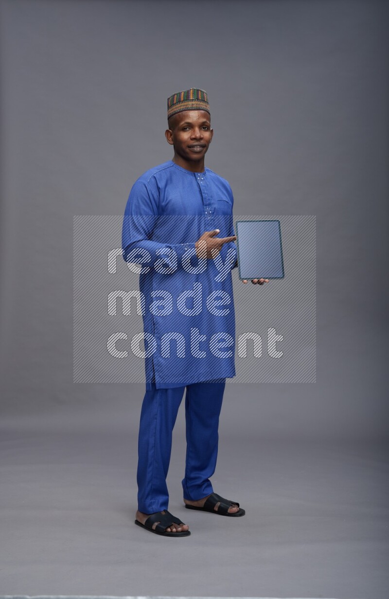 Man wearing Nigerian outfit standing showing tablet to camera on gray background