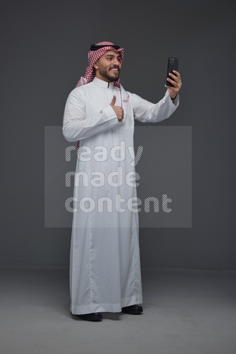 A Saudi man wearing Thobe and Shamgh taking selfie with his phone eye level on a gray background