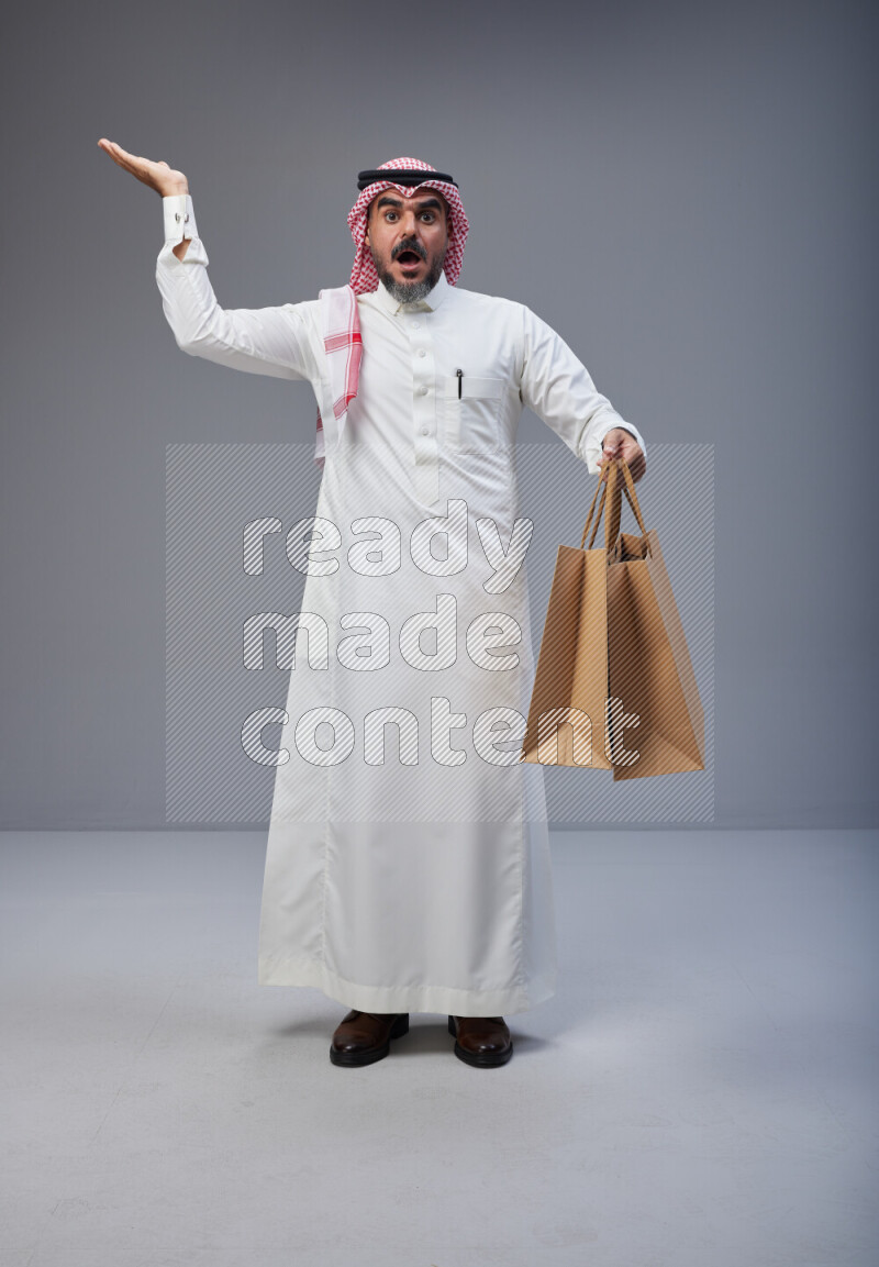 Saudi man Wearing Thob and red Shomag standing holding shopping bag on Gray background