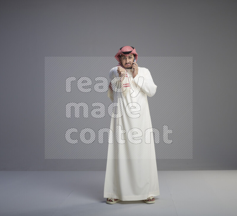 A Saudi man standing wearing thob and red shomag talking on phone on gray background