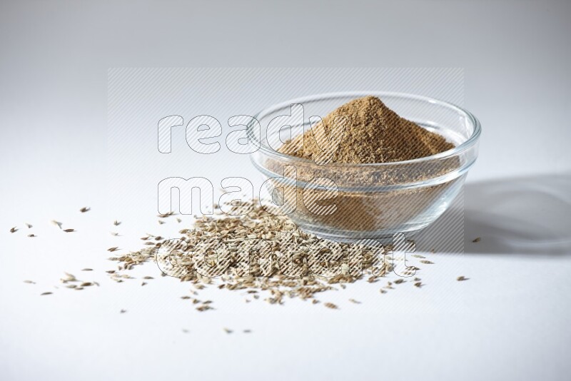 A glass bowl full of cumin powder with cumin seeds beneath it on white flooring