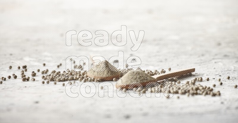 2 wooden spoons full of white pepper powder with white pepper beads on textured white flooring