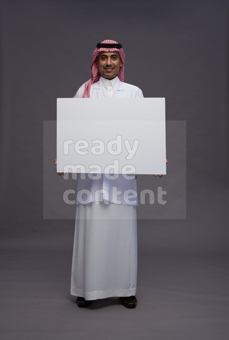 Saudi man wearing thob with lab coat and shomag with pocket employee badge standing holding board on gray background