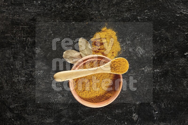 A wooden bowl and a wooden spoon full of turmeric powder on textured black flooring