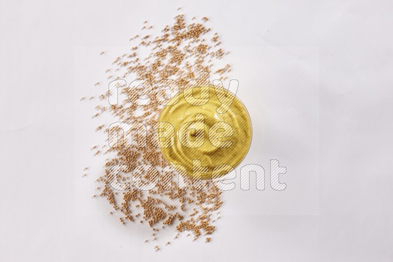 A glass bowl full of mustard paste with mustard seeds underneath on white flooring