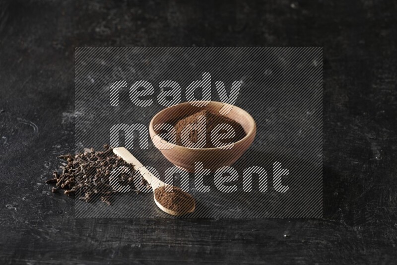 A wooden bowl and a wooden spoon full of cloves powder with spreaded cloves on a textured black flooring