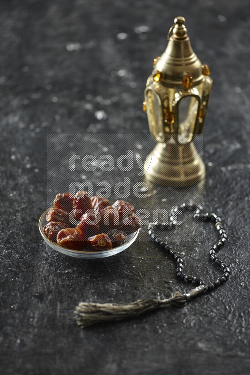 A golden lantern with different drinks, dates, nuts, prayer beads and quran on textured black background