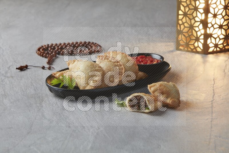 Five fried sambosas in an oval shaped black plate, beside a cut meat sambosa, a brown misbaha and a golden lantern on a gray background