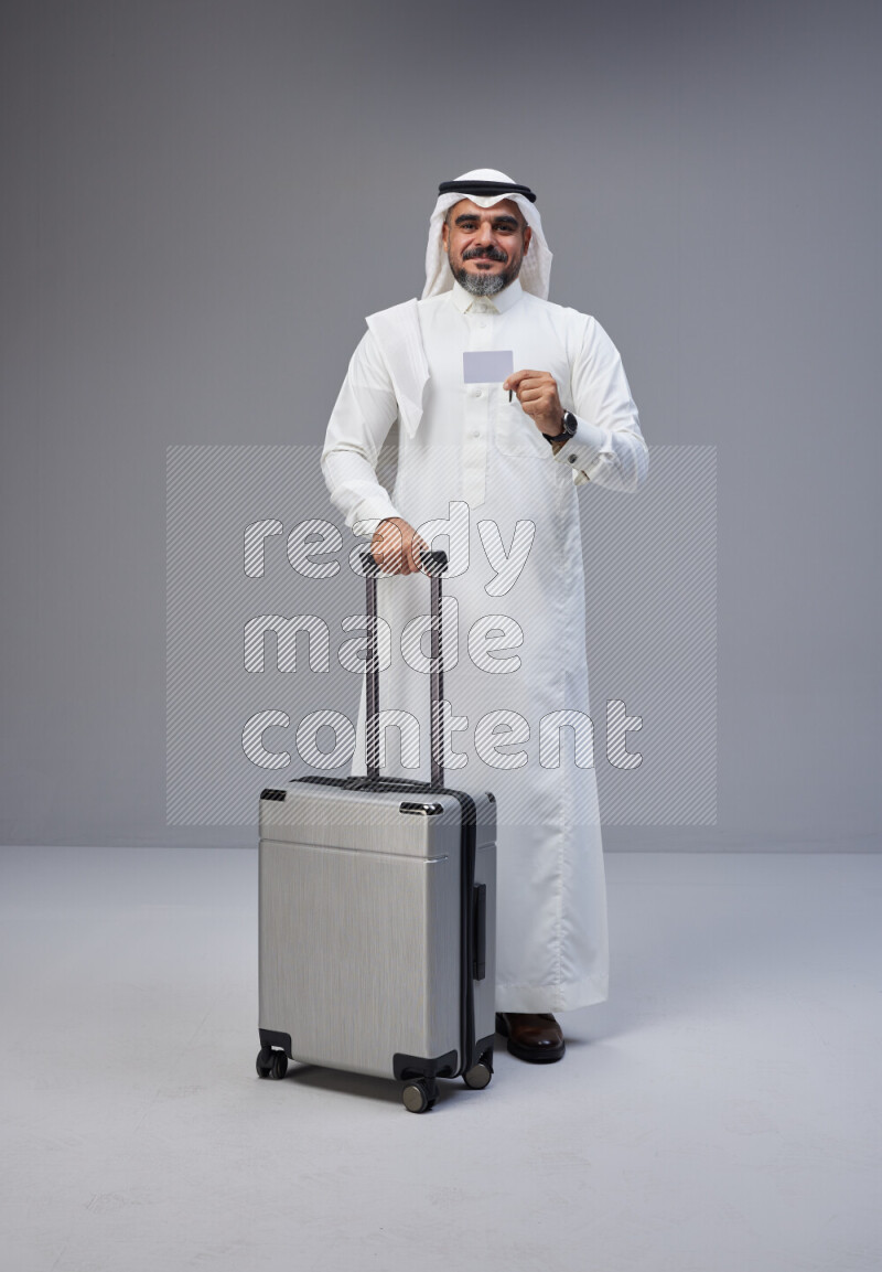 Saudi man wearing Thob and white Shomag standing holding Travel bag and ATM card on Gray background