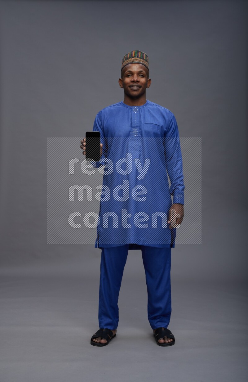Man wearing Nigerian outfit standing showing phone to camera on gray background