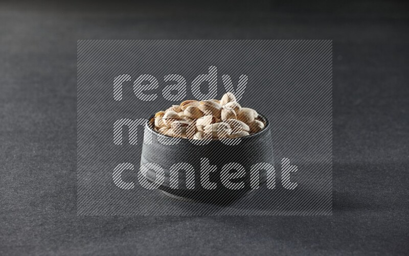 A black pottery bowl full of pistachios on a black background in different angles