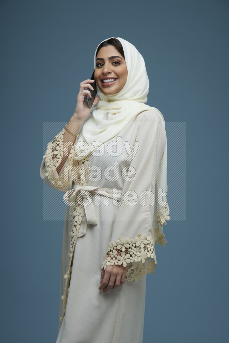 A Saudi woman having a call in a blue background wearing an off-white Abaya Hijab