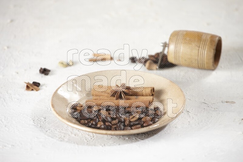 Beige plate full of coffee beans, cinnamon sticks and star anise with a coffee grinder, coffee beans, cinnamon pieces and cardamom next of it on white background