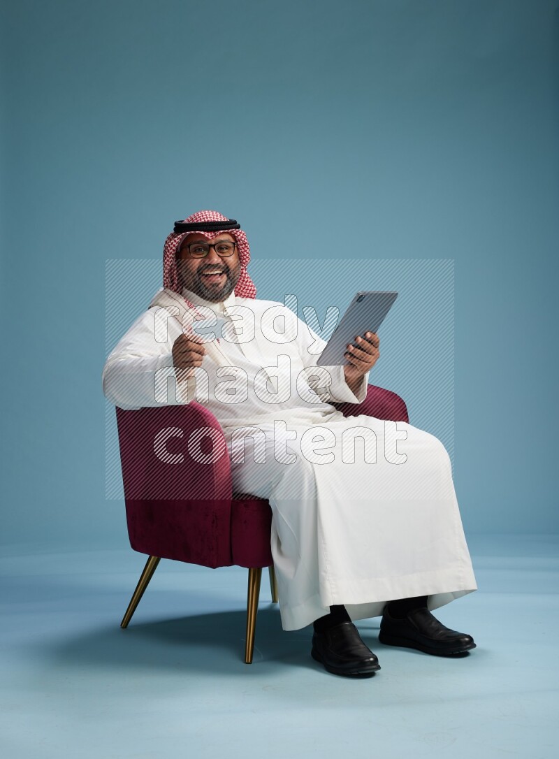 Saudi Man with shimag sitting on chair holding ATM card while working on tablet on blue background