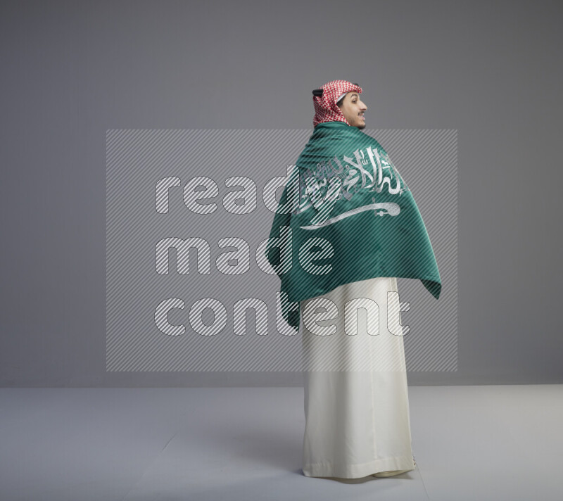 A Saudi man standing wearing thob and red shomag wrapping big saudi flag on gray background