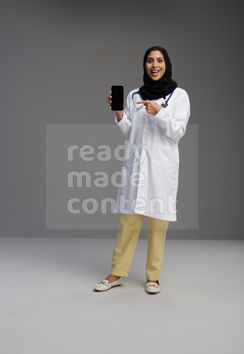 Saudi woman wearing lab coat with stethoscope standing showing phone to camera with sign in the back on Gray background