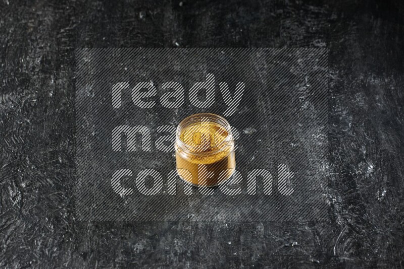 A glass jar full of turmeric powder on a textured black flooring