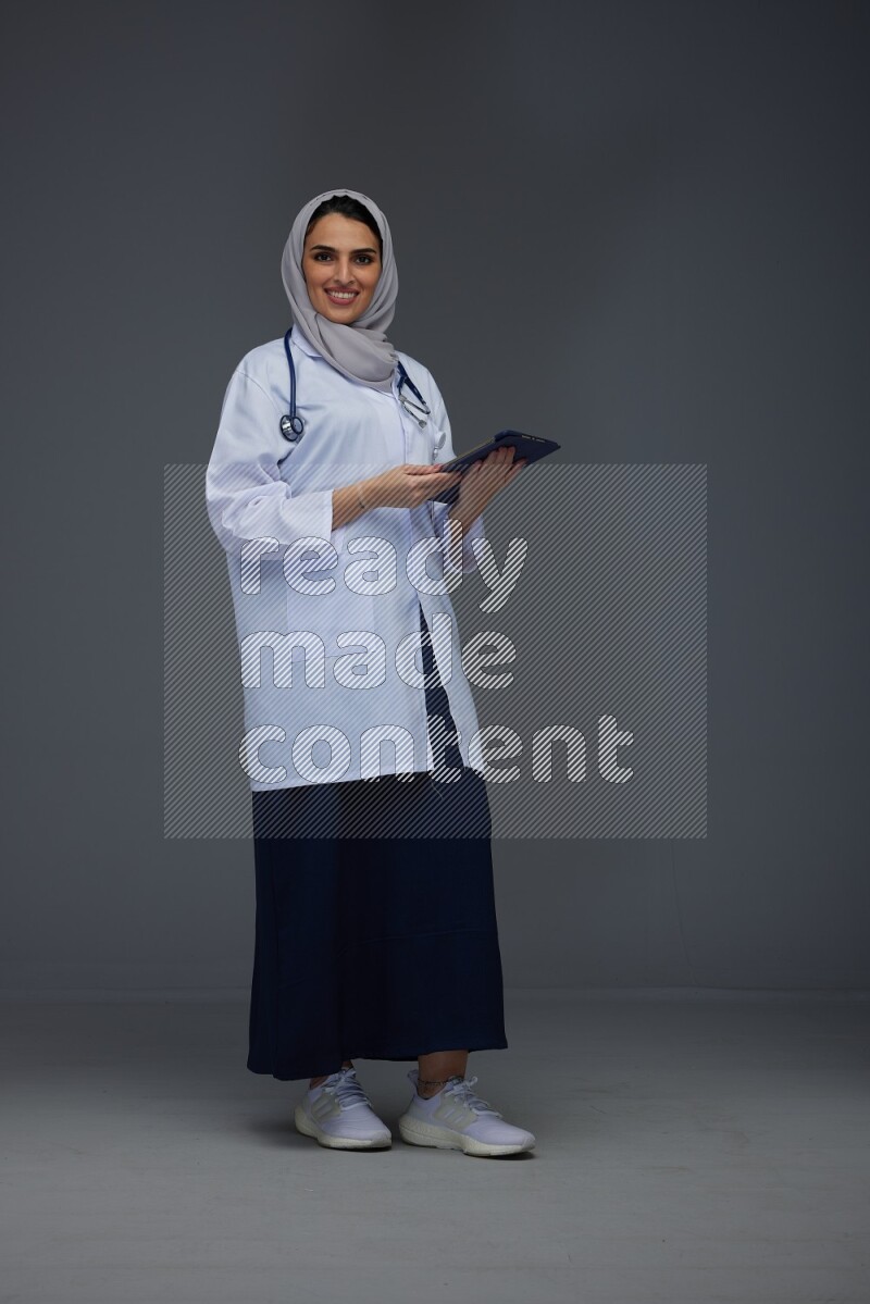 A Saudi doctor wearing a light gray head scarf standing and crossing her hands eye level on a grey background