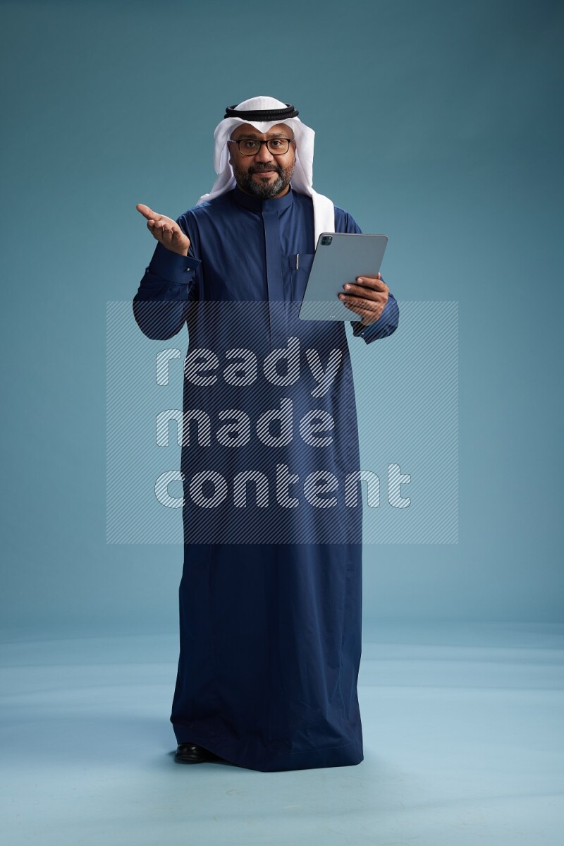 Saudi Man with shimag Standing holding ATM card while working on tablet on blue background