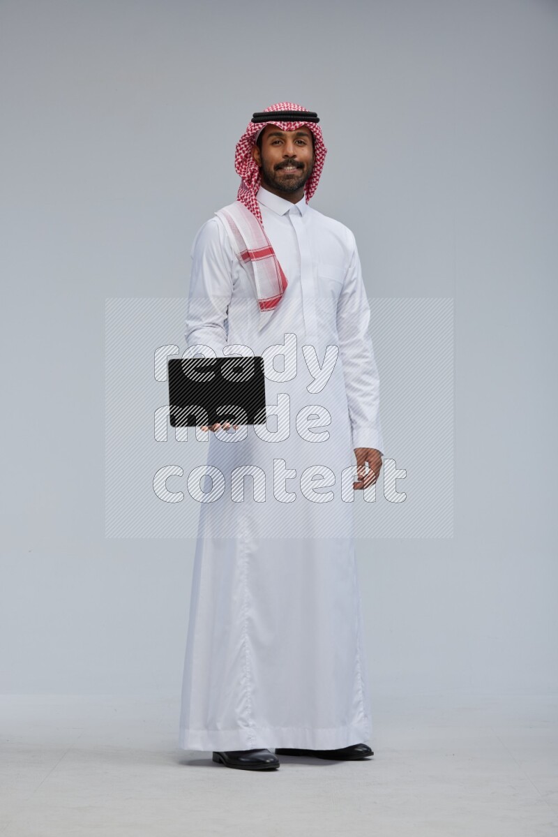 Saudi man Wearing Thob and shomag standing showing tablet to camera on Gray background