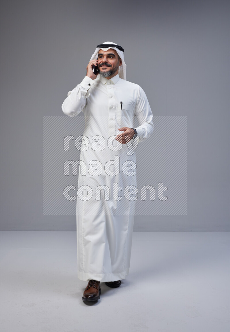 Saudi man Wearing Thob and white Shomag standing talking on phone on Gray background