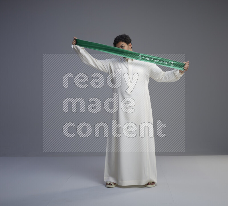 A Saudi man standing wearing thob holding Saudi flag scarf on gray background