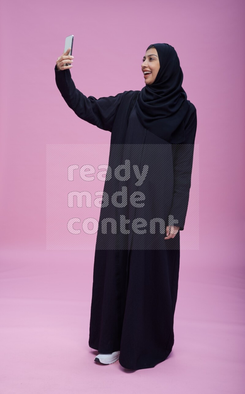 Saudi woman wearing Abaya standing taking selfie on pink background