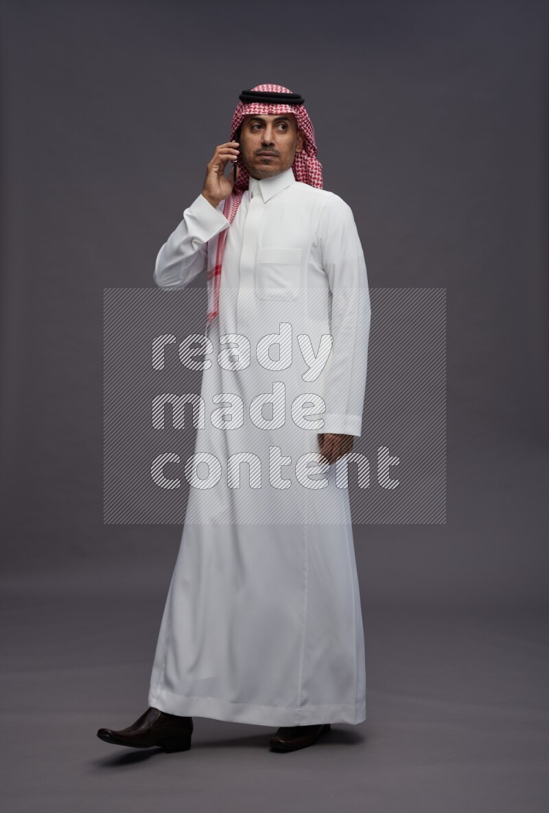Saudi man wearing thob and shomag standing talking on phone on gray background