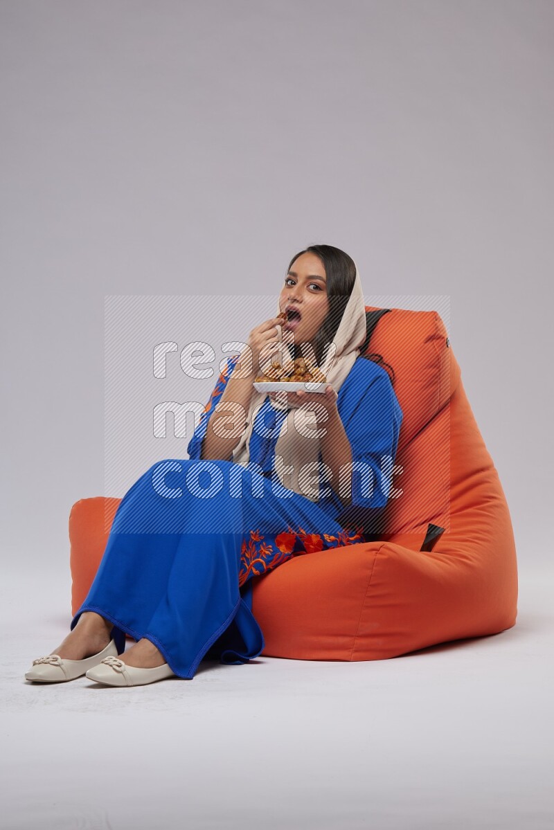 A Woman sitting on an orange beanbag wearing Jalabeya holding a plate of dates