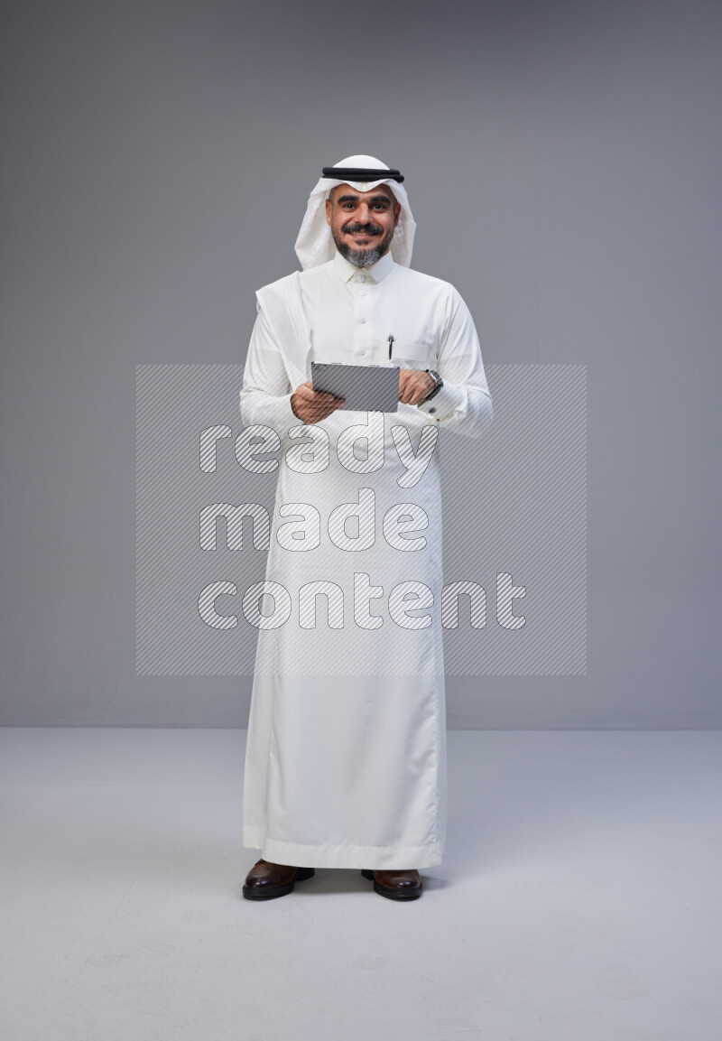 Saudi man Wearing Thob and white Shomag standing working on tablet on Gray background