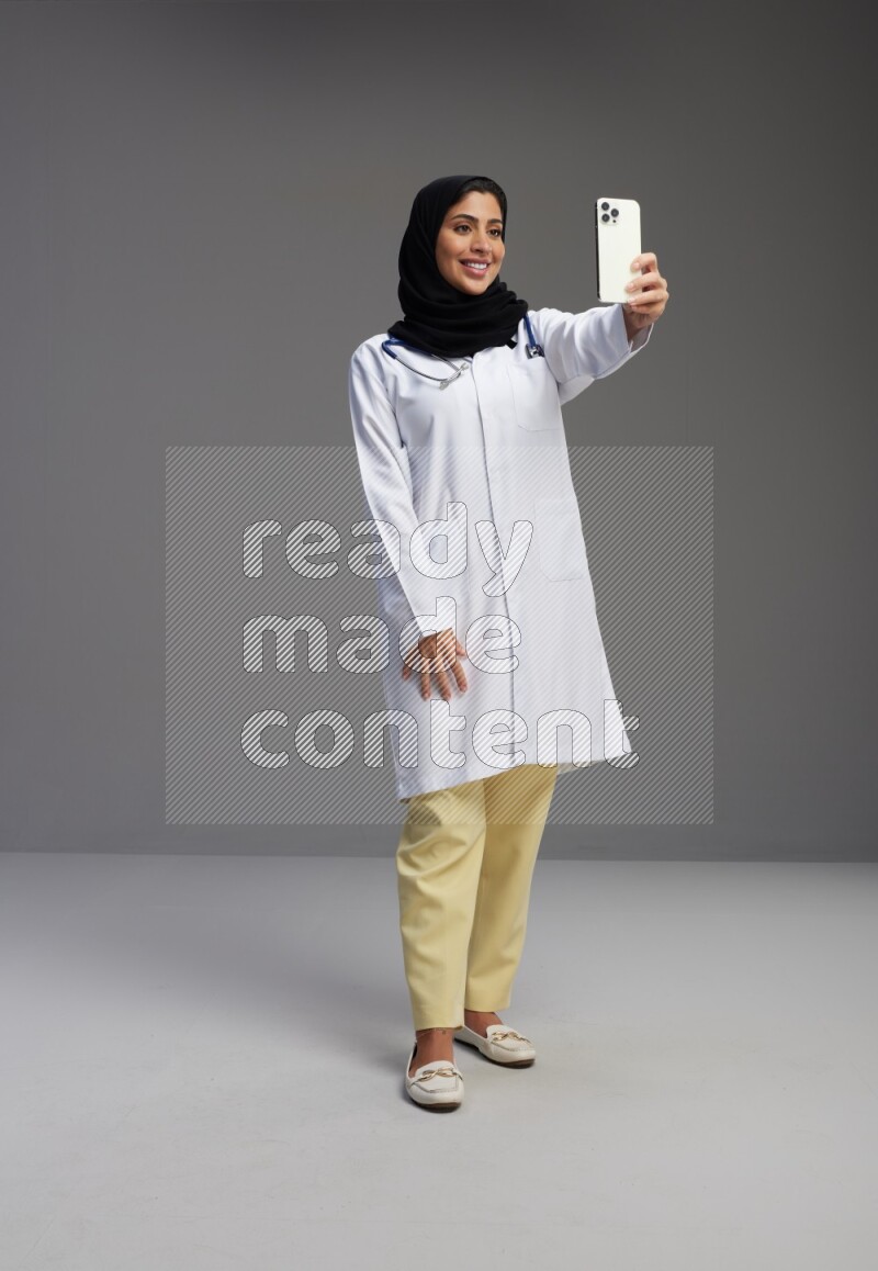 Saudi woman wearing lab coat with stethoscope standing taking selfie on Gray background