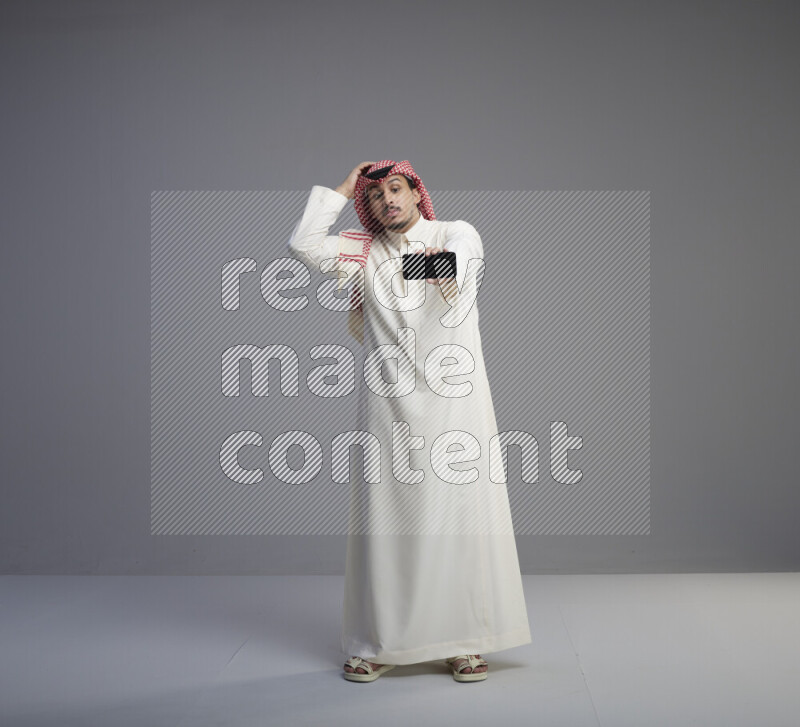A Saudi man standing wearing thob and red shomag showing phone to camera on gray background