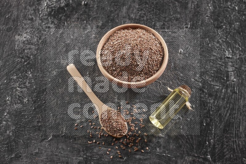 A wooden bowl and spoon full of flaxseeds and a glass bottle of flaxseeds oil on a textured black flooring