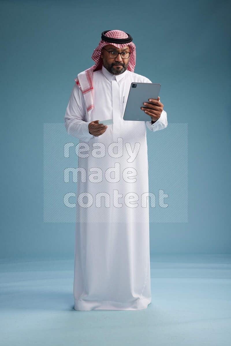 Saudi Man with shimag Standing holding ATM card while working on tablet on blue background