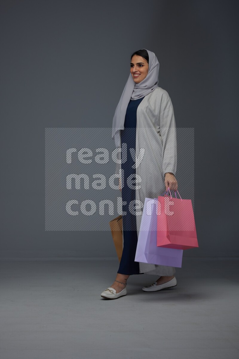 A Saudi woman wearing a light gray Abaya and head scarf standing and holding shopping bags on a grey background