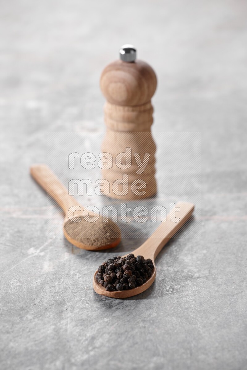 2 wooden spoons, one full of black pepper powder and the other full of black pepper beads and wooden grinder on a marble flooring
