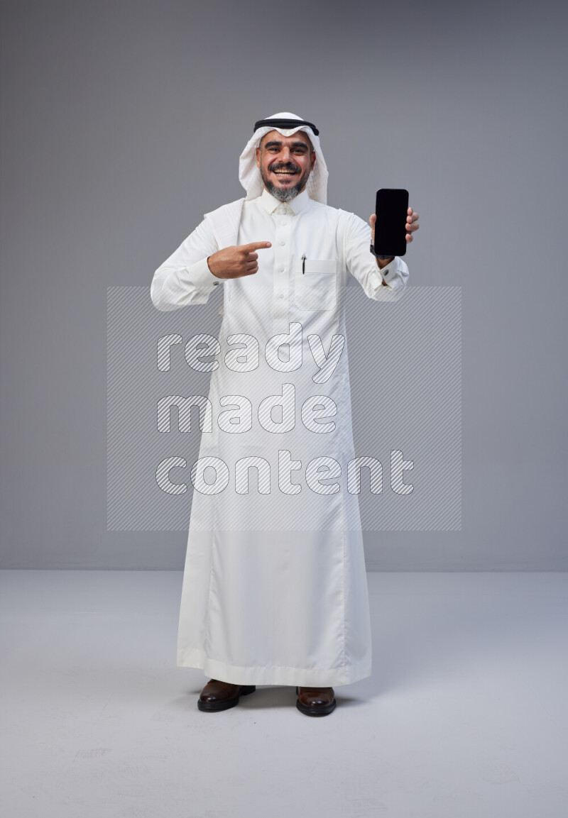 Saudi man Wearing Thob and white Shomag standing showing phone to camera on Gray background