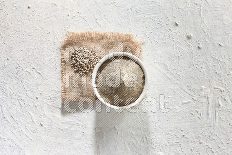 White pottery bowl full of white pepper powder set on a burlap piece of fabric with pepper beads and wooden pepper grinder on textured white flooring