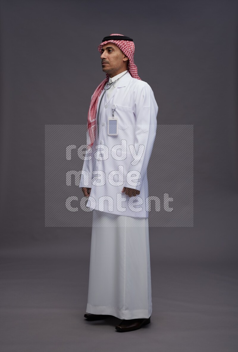 Saudi man wearing thob with lab coat and shomag with pocket employee badge with stethoscope standing interacting with the camera on gray background