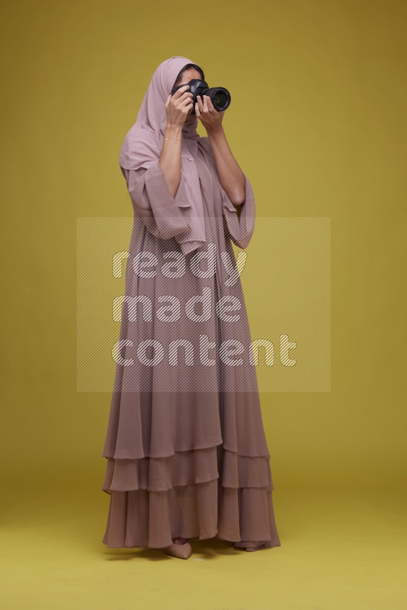 A woman Holding a Camera on a Yellow Background wearing Brown Abaya with Hijab