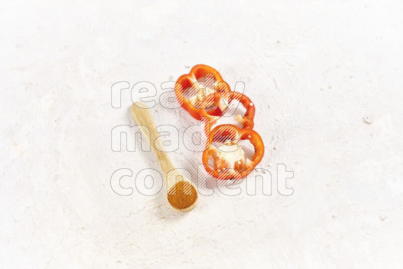 A wooden spoon full of ground paprika powder with red bell pepper slices beside it on white background