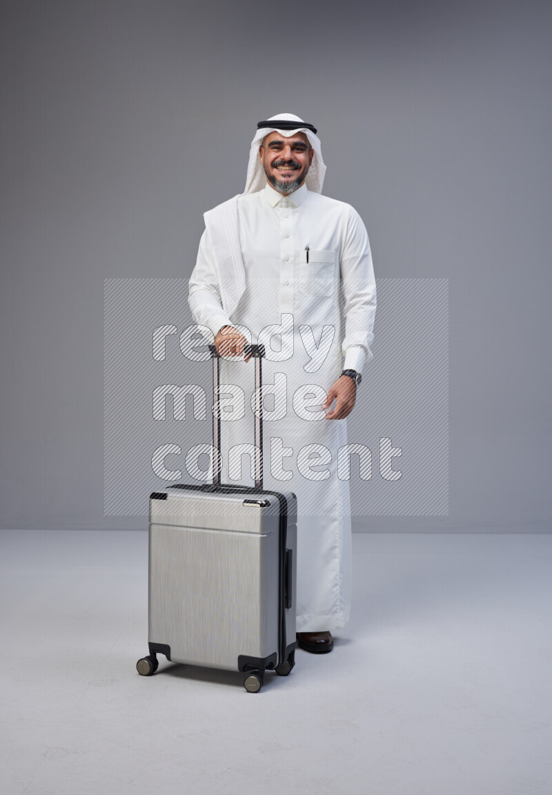 Saudi man wearing Thob and white Shomag standing holding Travel bag on Gray background